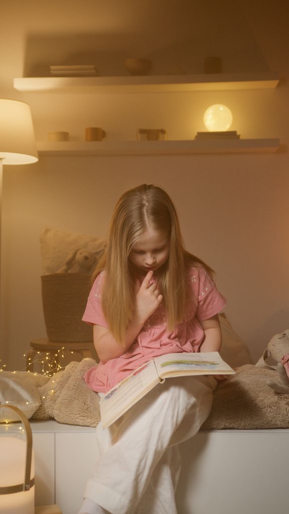A young girl in a pink dress reads a storybook in a cozy bedroom setting with warm lighting.